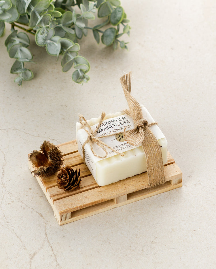 Steinhäger Männerseife auf eine Holz Palette. Als Dekoration wird ein kleiner Tannenzapfen und eine offene Buchecker angebracht und die Seife mit einem Schleifenband befestigt. Mit Deko